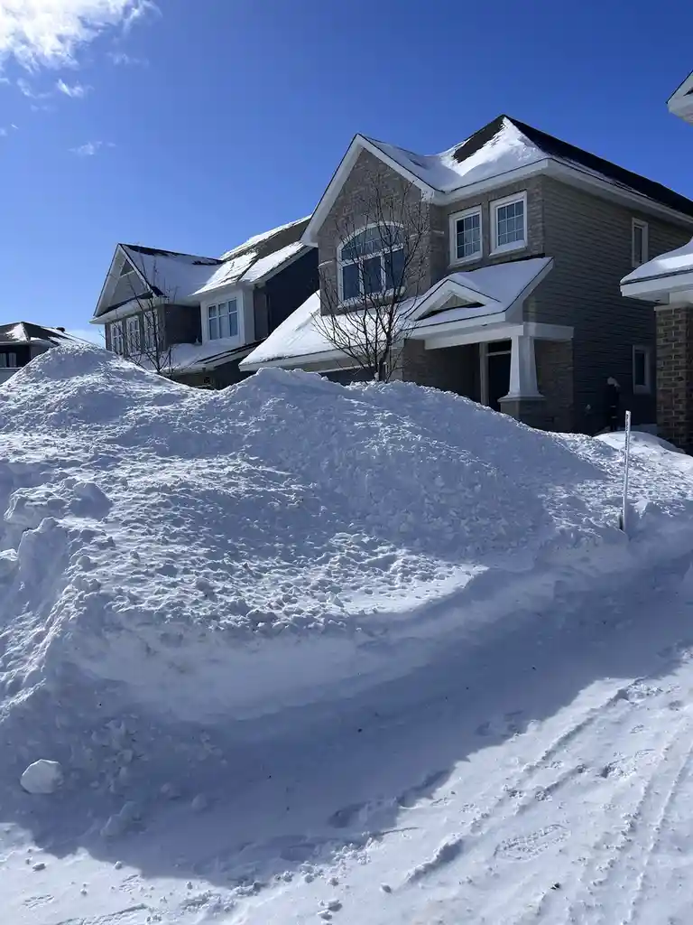 A large, uneven mound of snow dominates the foreground of a residential street, with a modern two-story house visible behind…