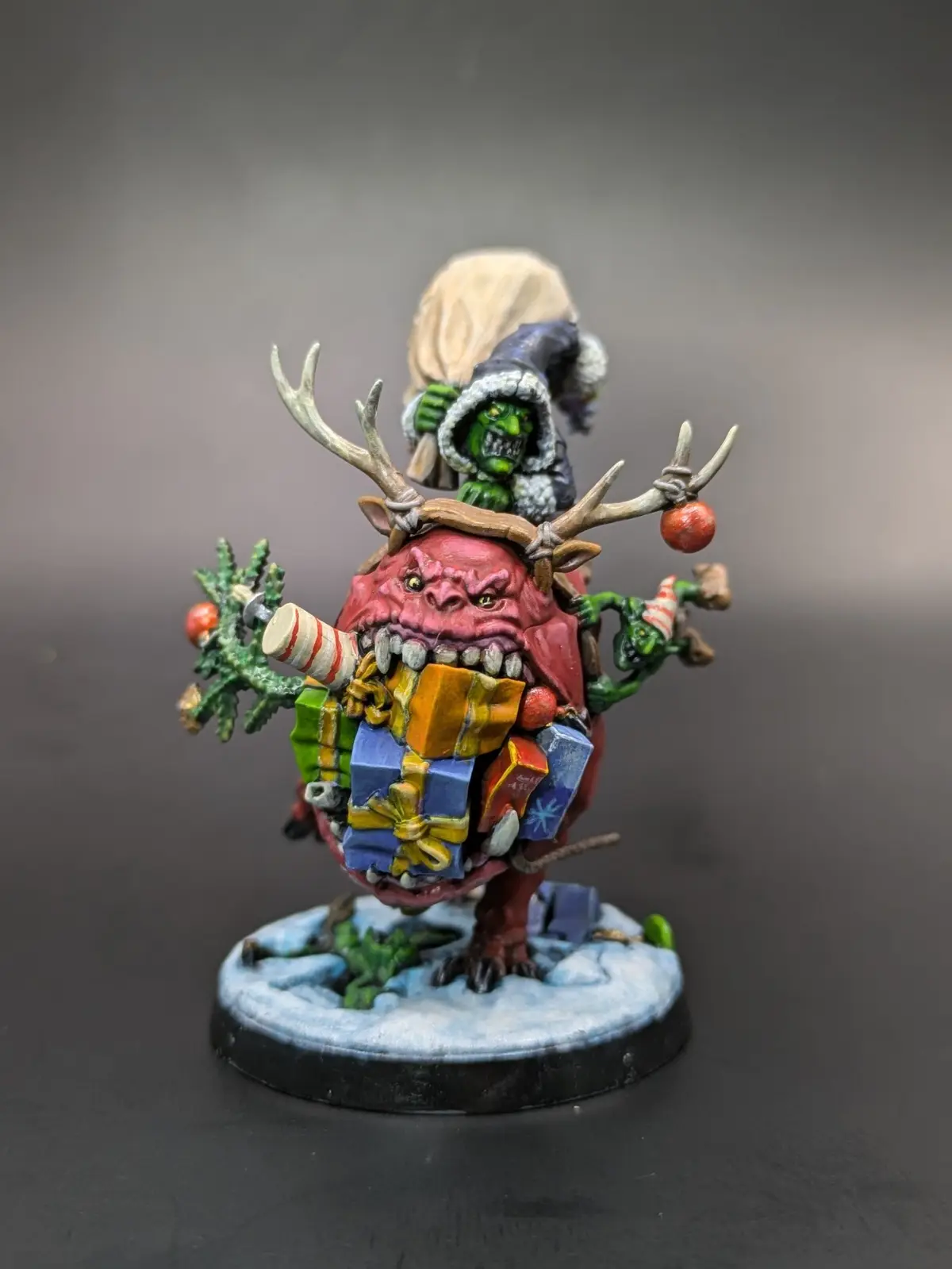 A painted miniature depicts a green-skinned orc wearing a winter hat and carrying a sack of gifts, riding atop a red, horned…