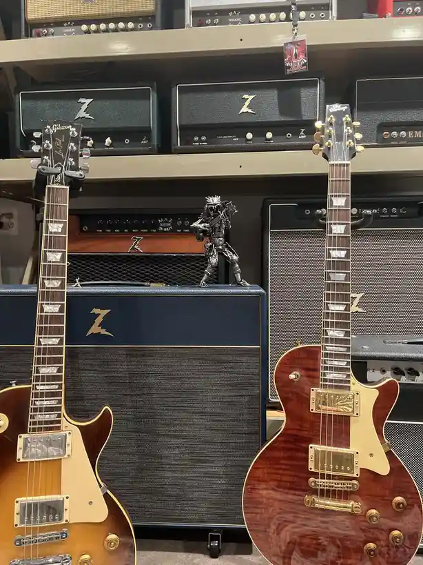 Two electric guitars stand in front of a wall of Z-brand amplifiers, with a small metal figurine of a guitarist perched on…