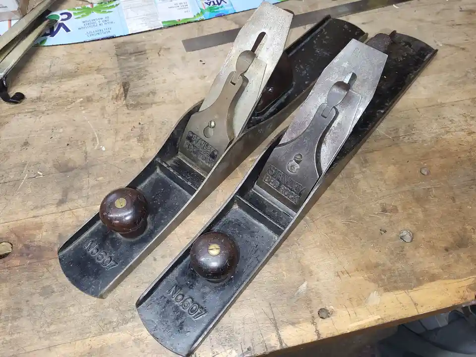 Two identical Stanley Bedrock No. 607 hand planes lie side by side on a well-worn wooden workbench, their metal bodies…