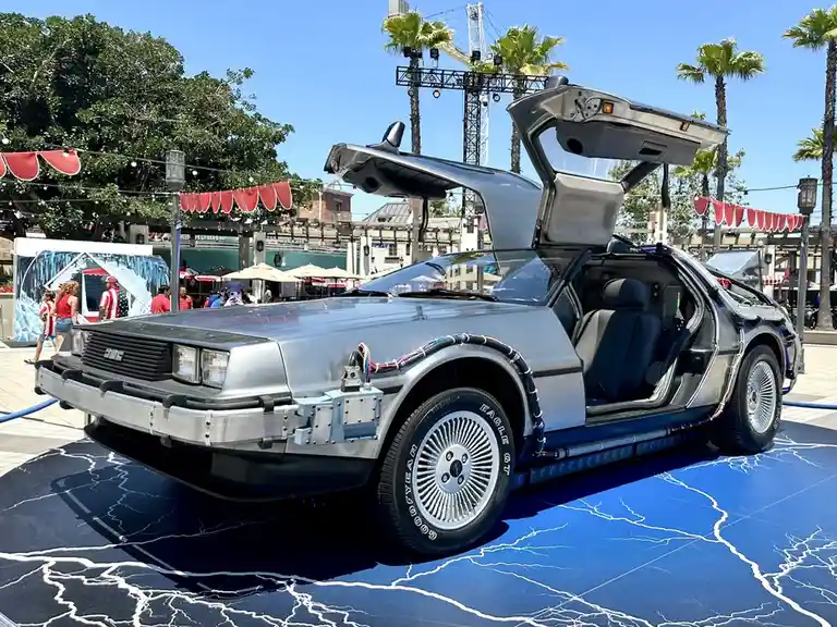 A silver DeLorean car with its gull-wing doors open, parked on a blue floor with white lightning bolt patterns. The car has…