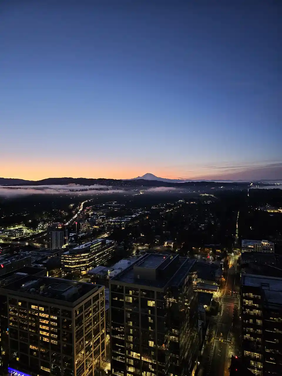 A high-angle cityscape at dusk with illuminated buildings, a fog-covered valley, and a snow-capped mountain on the horizon…