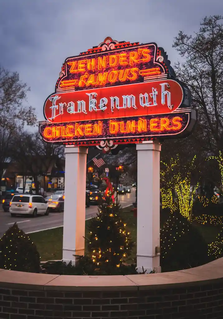 A large, ornate, illuminated neon sign for 'Zehnder's Famous Frankenmuth Chicken Dinners' stands at dusk, supported by two…