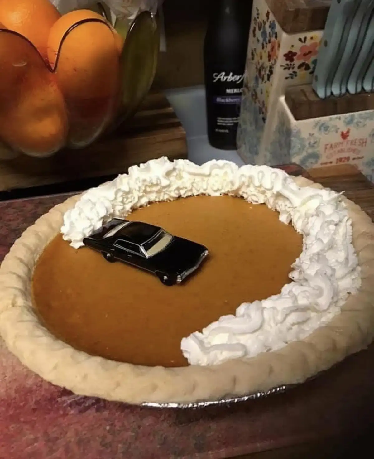 A close-up photo shows a pumpkin pie with whipped cream around the edge, and a small black toy car placed in the center of…