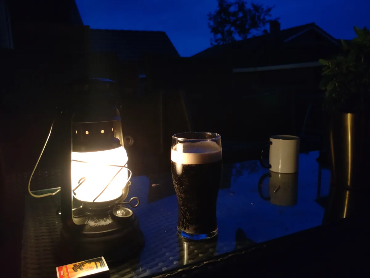 A dimly lit outdoor table at dusk features a glowing lantern, a glass of dark beer with a foamy head, and a white mug. The…
