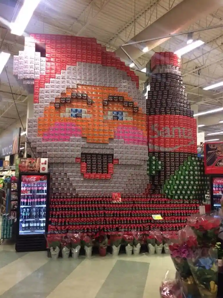 A large, pixelated Santa Claus figure constructed entirely from stacked Coca-Cola cans and bottles is displayed inside a…