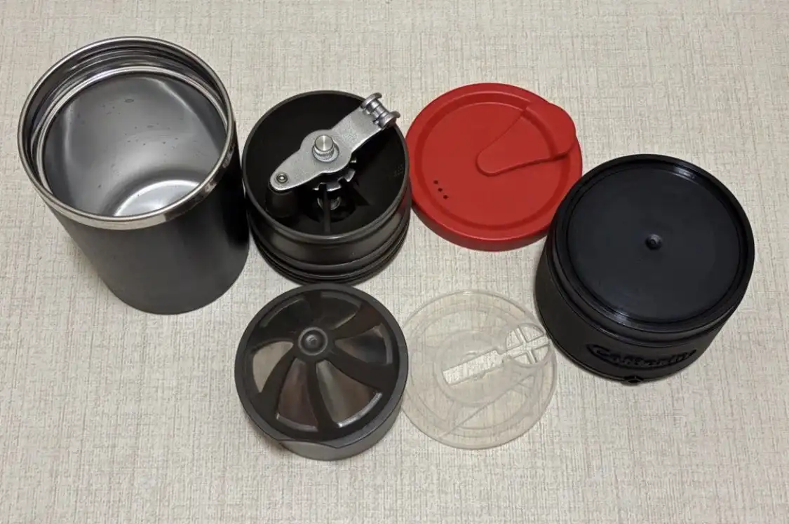 A disassembled coffee grinder with multiple parts laid out on a beige fabric surface, including a metal cup, grinding…