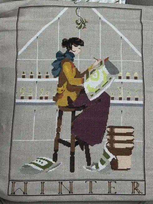 A cross-stitch embroidery depicts a woman in a greenhouse, holding a large, stylized green eye, with shelves of potted…