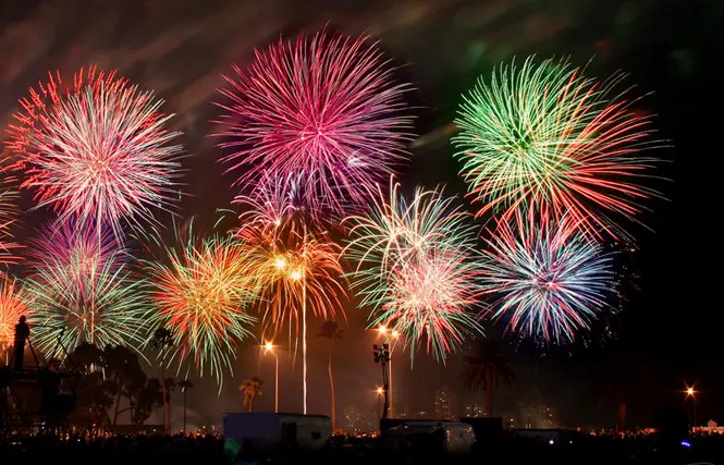 A nighttime photograph shows multiple colorful fireworks exploding in the sky above a crowd and palm trees, with the word…