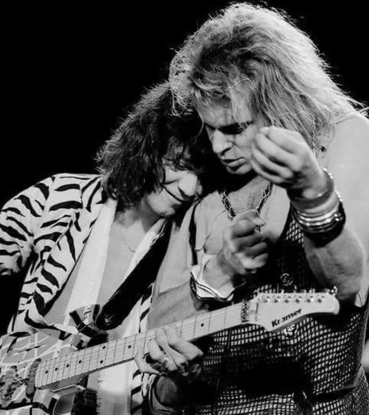 A black-and-white photo of two men on stage, one with long curly hair in a zebra-print jacket playing a Kramer guitar, the…