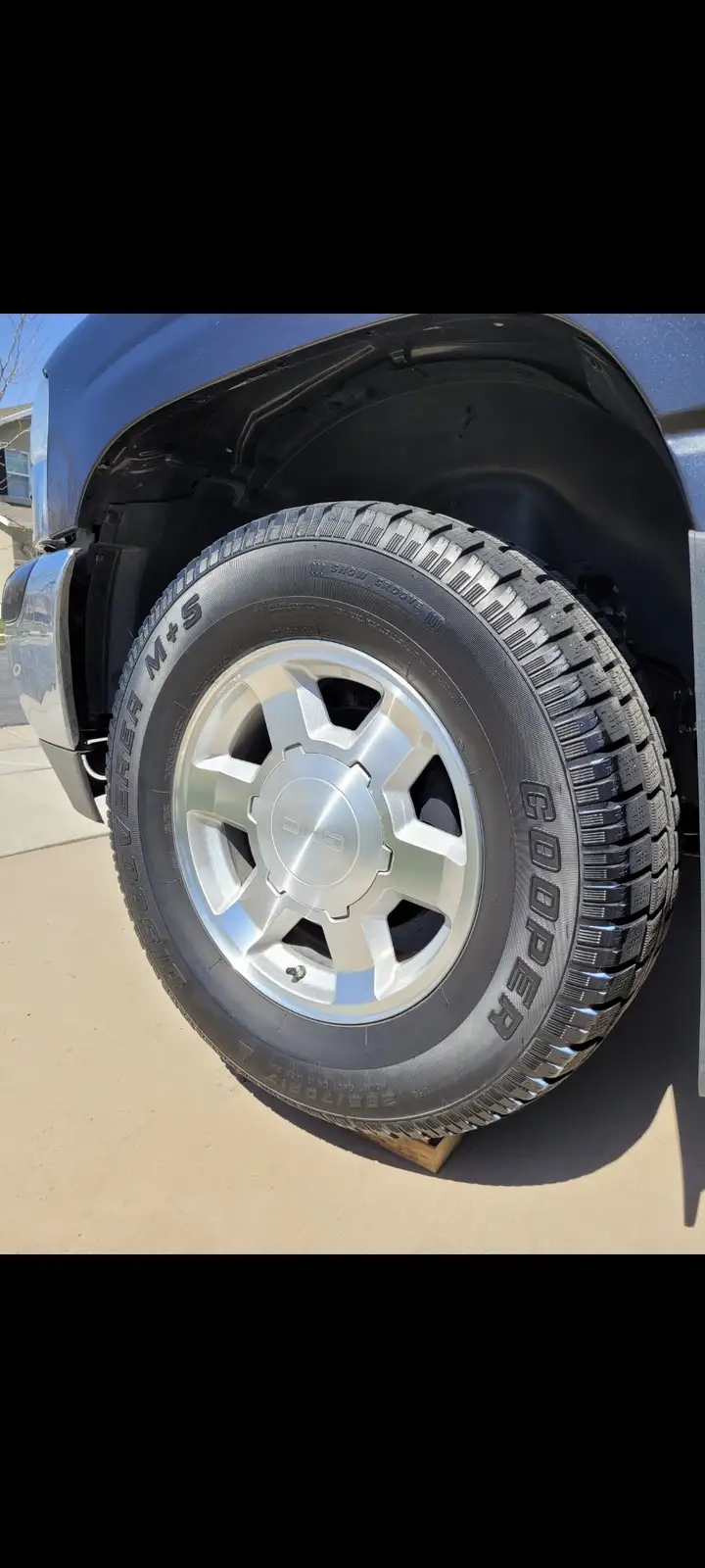 A close-up photo shows a GMC truck's wheel with a Cooper Discoverer M+S tire, resting on a wooden block on a driveway. The…
