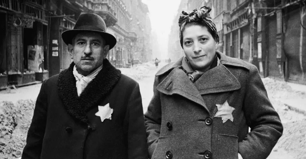 A black-and-white historical photograph shows a man and a woman standing on a snowy city street, both wearing heavy coats…