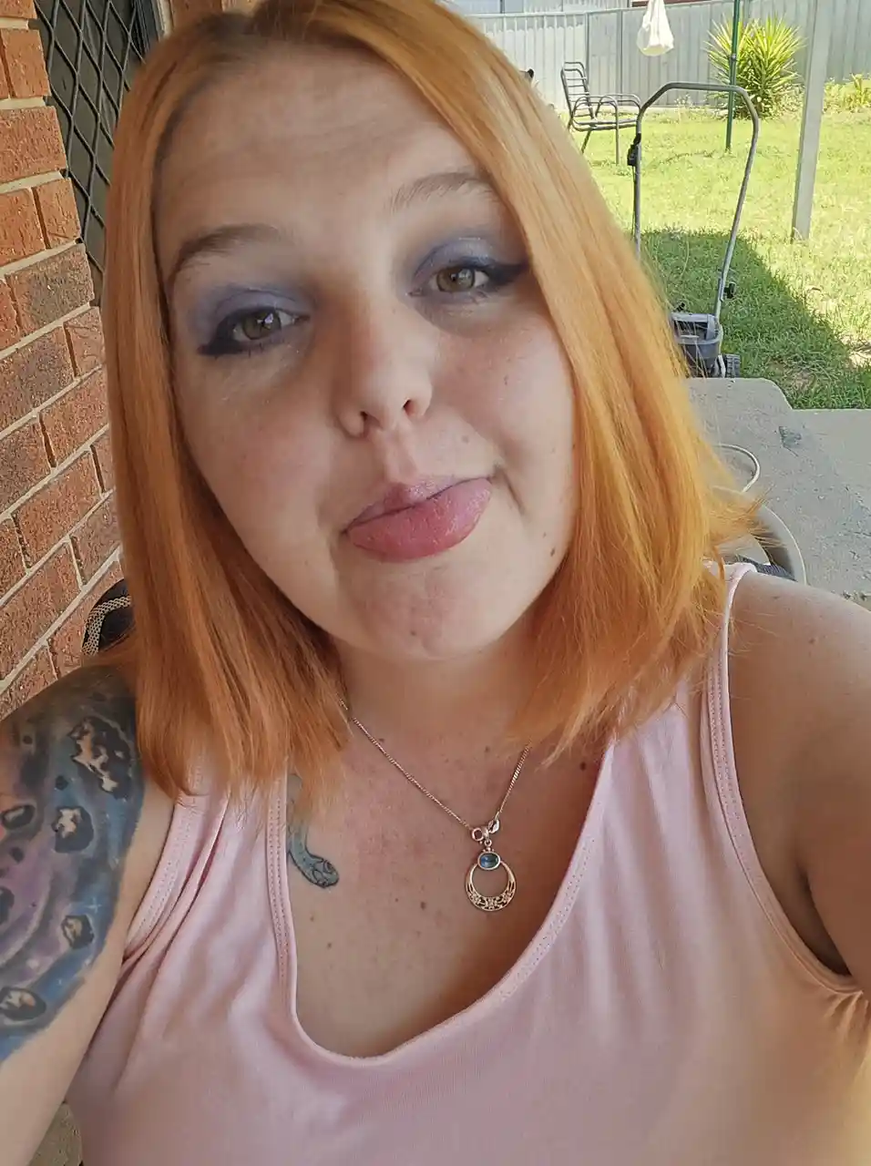 A selfie of a woman with orange hair and purple eyeshadow sticking her tongue out playfully while wearing a pink tank top…