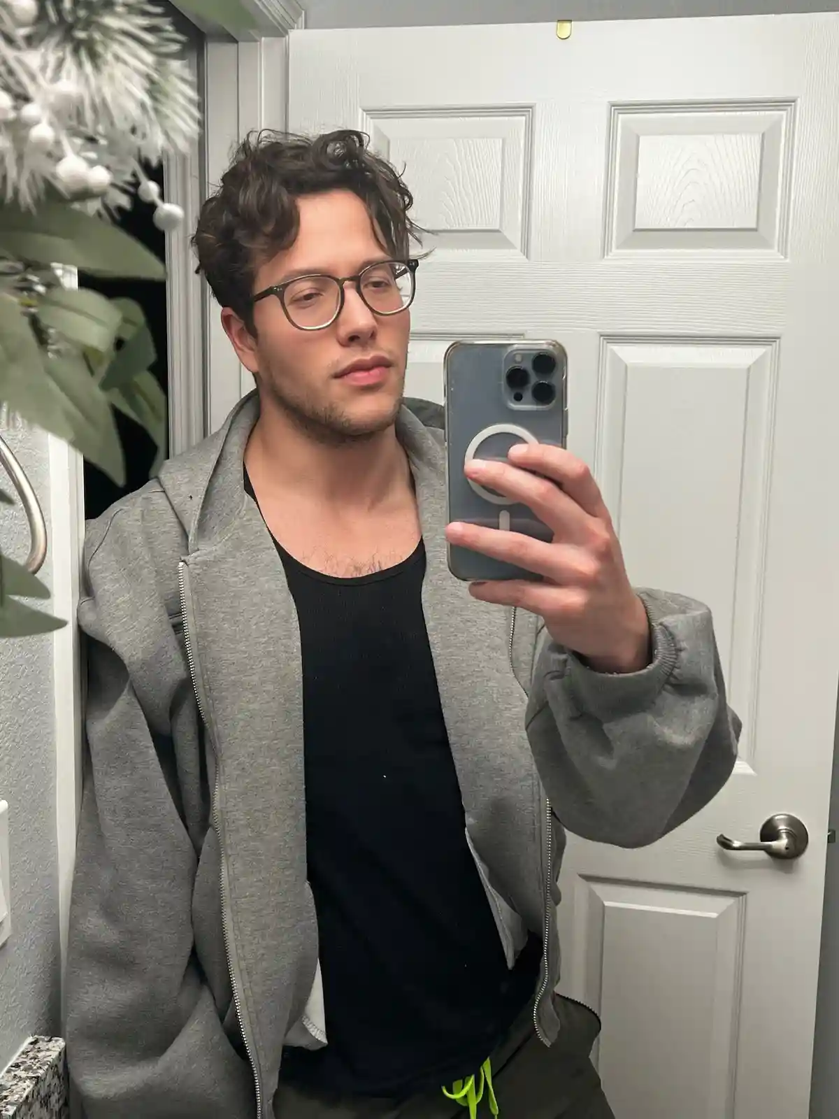 A young man with curly hair and glasses takes a mirror selfie holding a smartphone, wearing a grey zip-up hoodie over a…