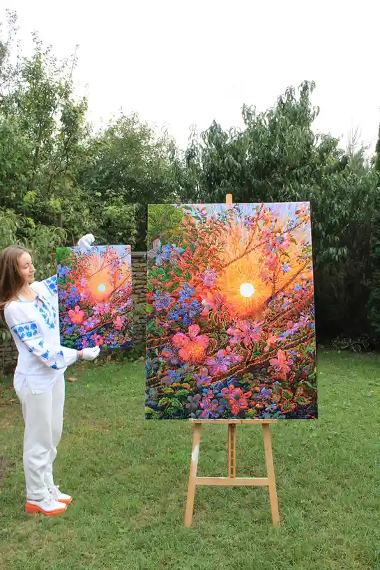 A woman in a white embroidered shirt and white gloves stands outdoors holding a small painting identical to a larger one on…