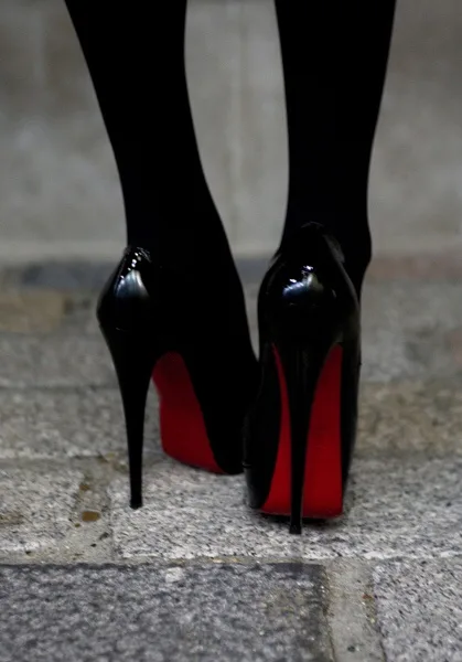A close-up shot of a person's feet wearing glossy black stiletto heels with iconic red soles, standing on a grey stone…
