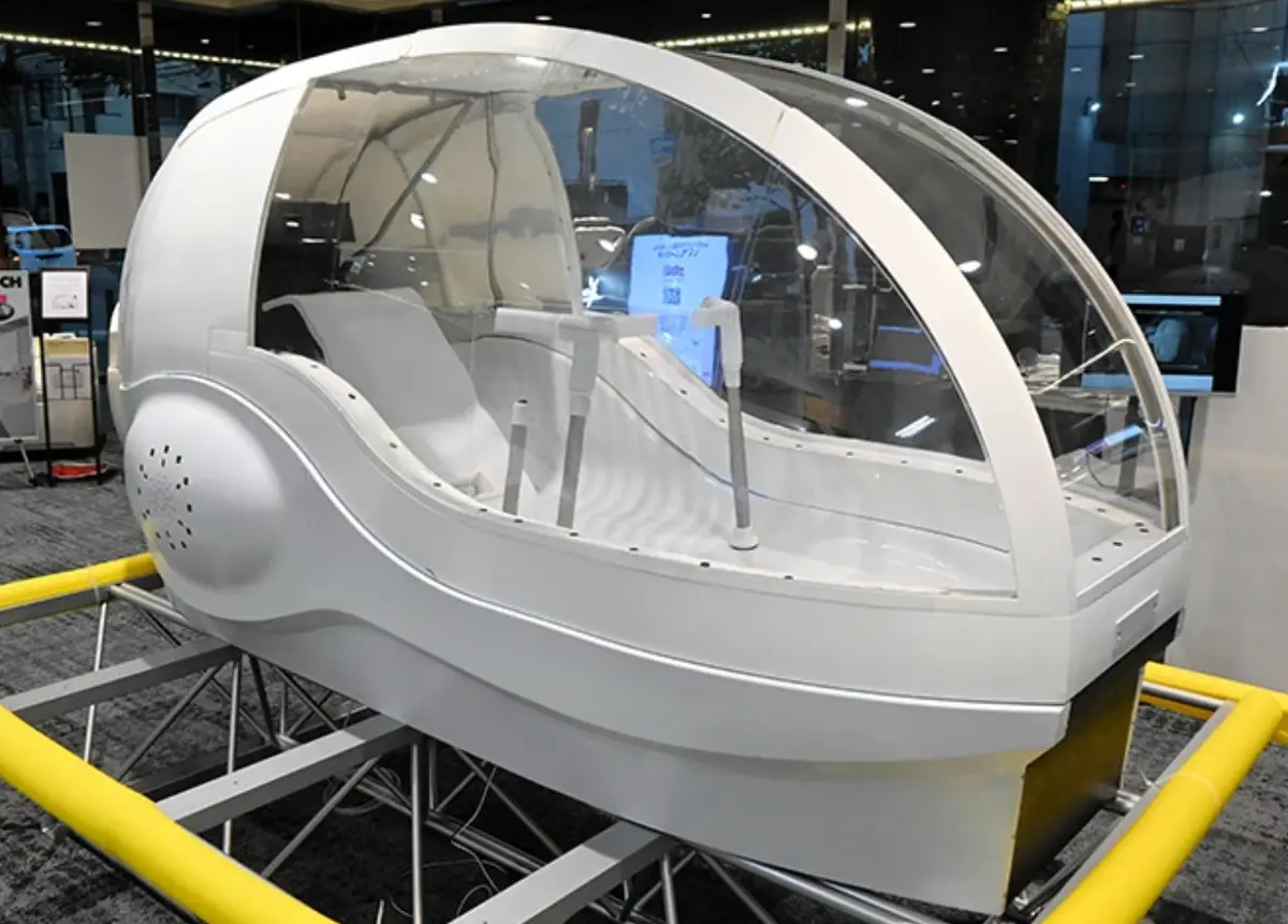 A sleek, white, futuristic-looking pod with a curved glass dome and interior seating is mounted on a metal frame with yellow…