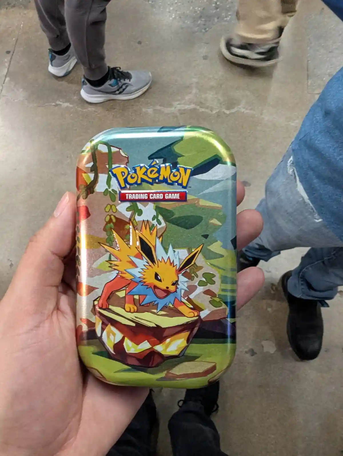 A hand holds a shiny Pokémon Trading Card Game tin featuring the Pokémon Jolteon on its cover, standing atop a large…