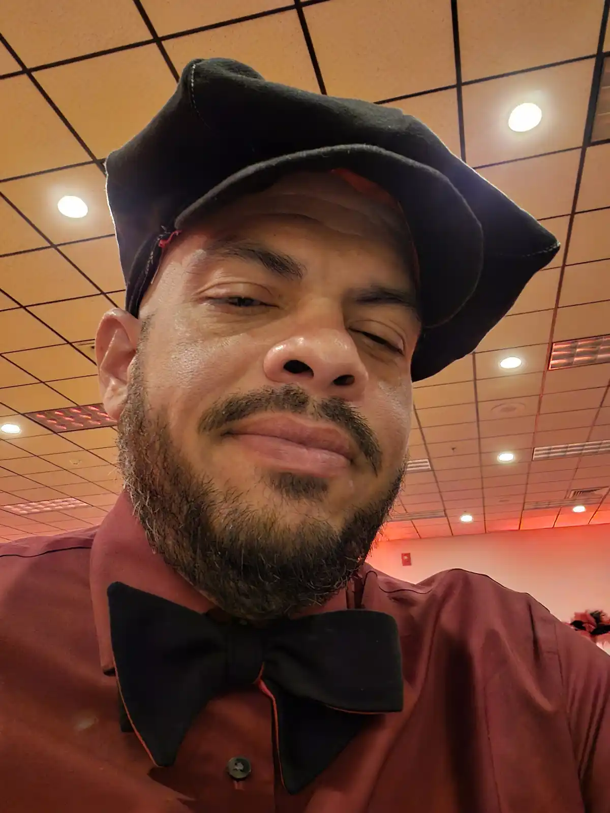 A man with a beard and mustache wears a black flat cap and a black bow tie over a red shirt, looking at the camera with his…