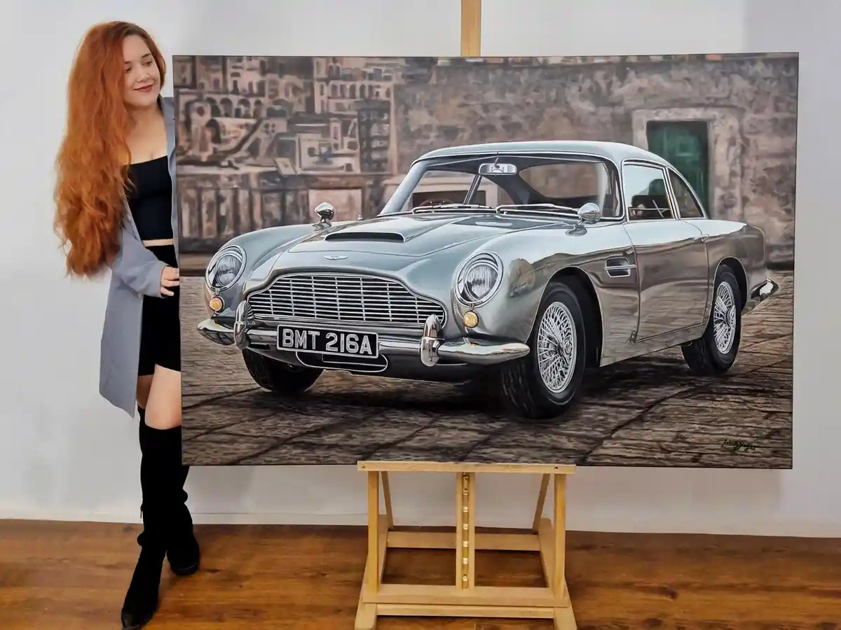 A woman with long red hair stands beside a large painting of a silver Aston Martin DB5, which is displayed on a wooden…