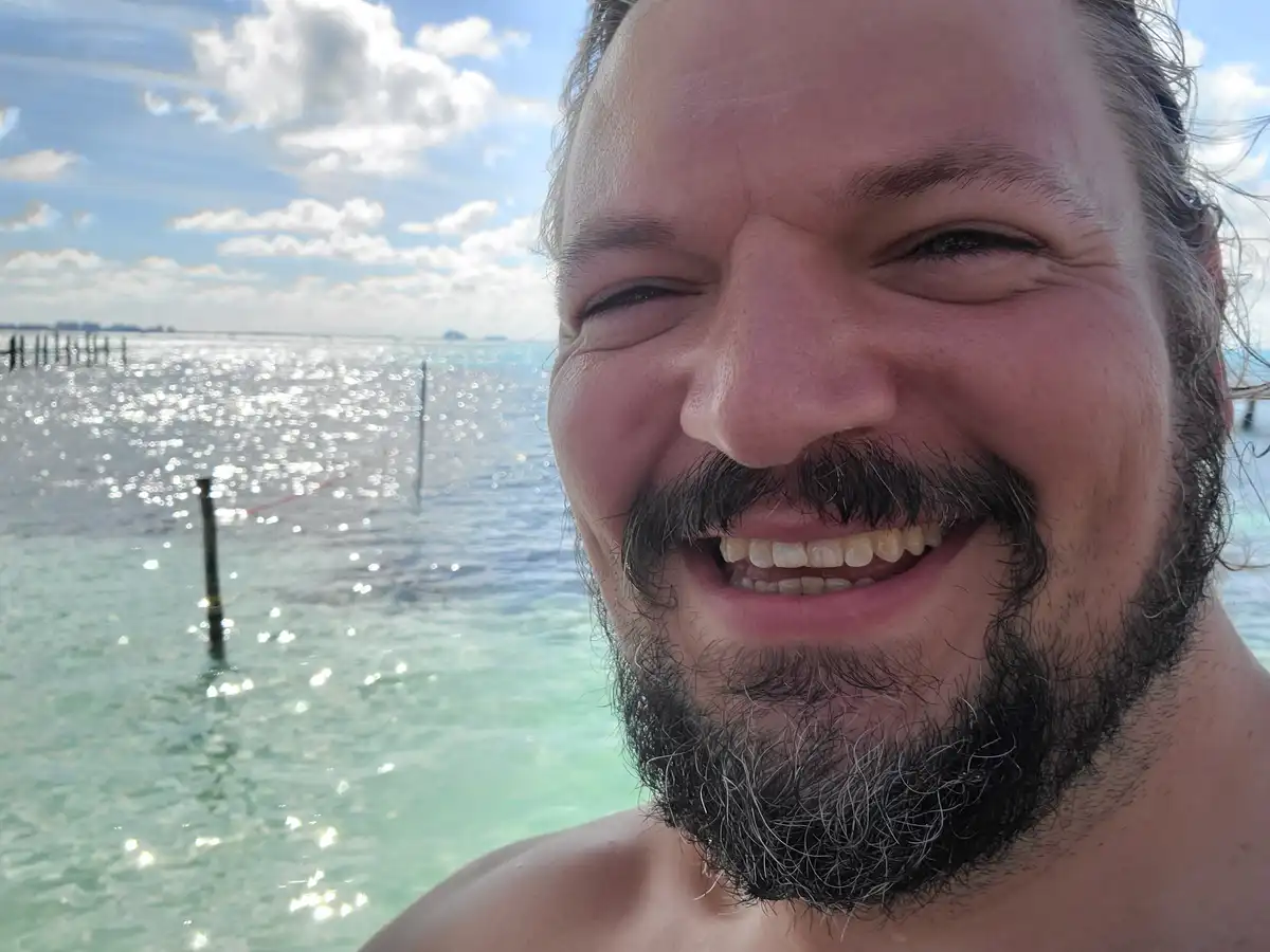 A man with a beard and mustache smiles broadly at the camera while standing in clear, sparkling turquoise water under a…