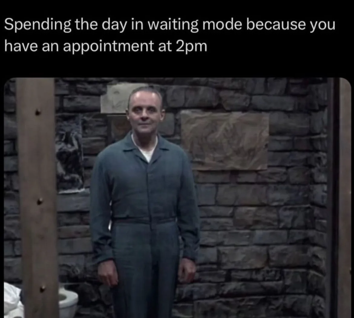A man in a grey jumpsuit stands motionless in front of a stone wall, looking directly at the camera with a blank, patient…