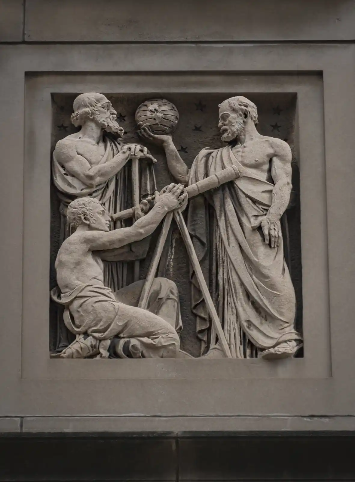 A stone relief depicts three classical figures: two standing men holding a globe and a telescope, and a kneeling man…