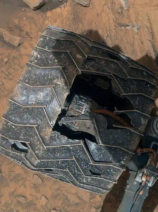 a close-up image of a damaged rover wheel on mars, showing a large hole in the tread exposing internal mechanical parts and…