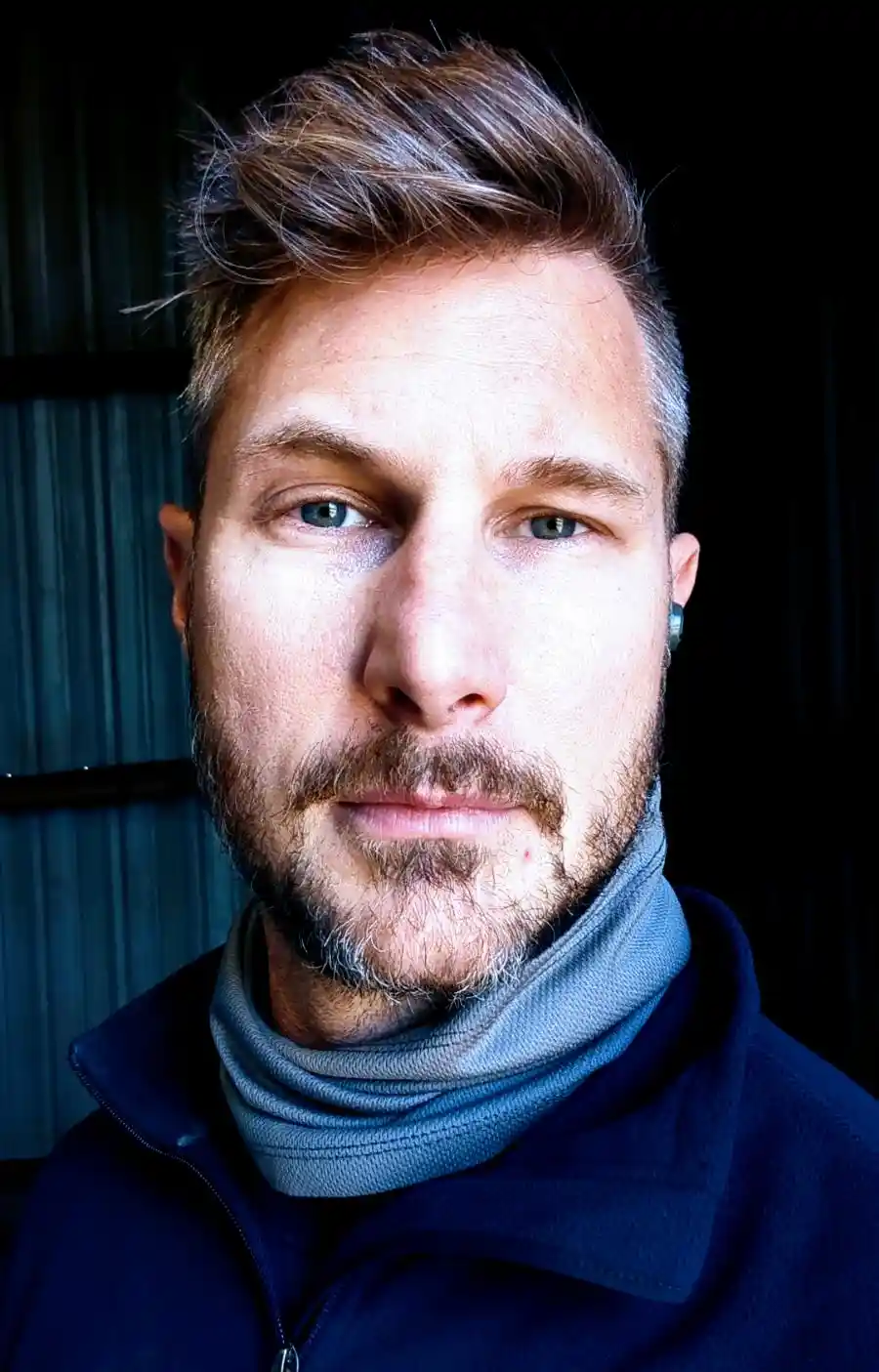 A man with styled brown hair, a beard, and a serious expression looks directly at the camera. He wears a blue jacket and a…