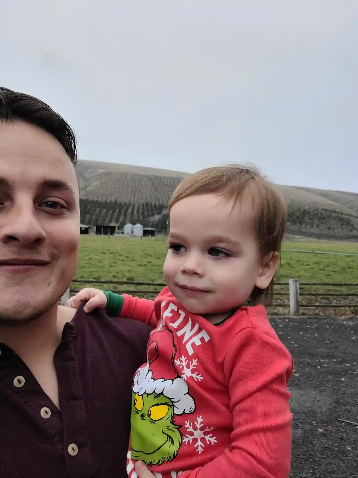 A man takes a selfie holding a toddler who is wearing a red holiday shirt featuring the Grinch and the word 'FINE' in large…