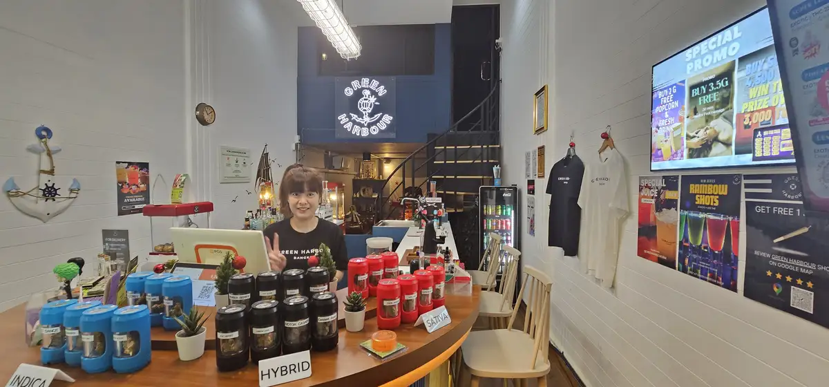 A smiling woman stands behind a cannabis dispensary counter labeled 'GREEN HARBOUR', surrounded by jars of cannabis strains…