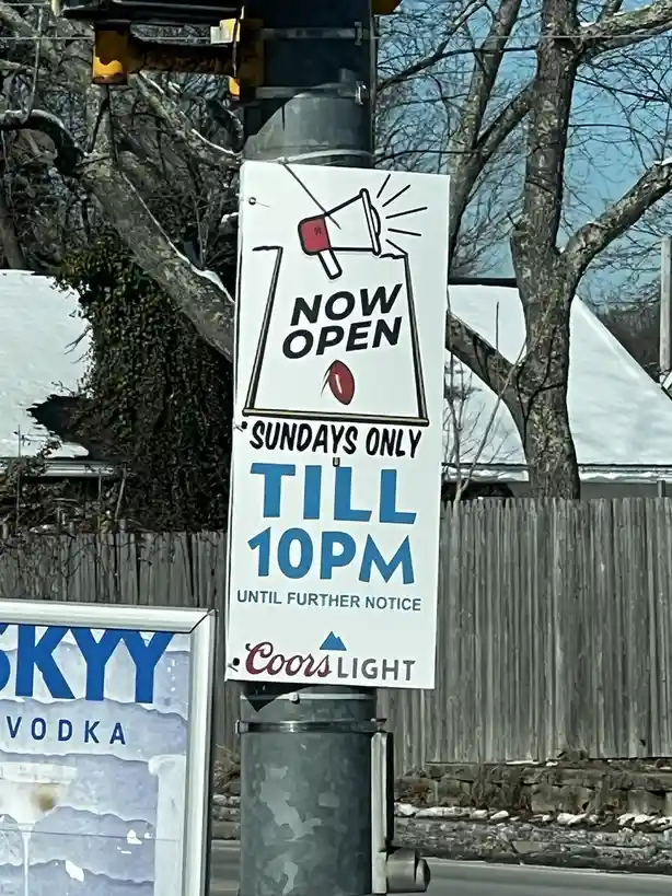 A white vertical sign on a pole advertises a business now open Sundays only until 10pm, featuring a megaphone graphic and a…
