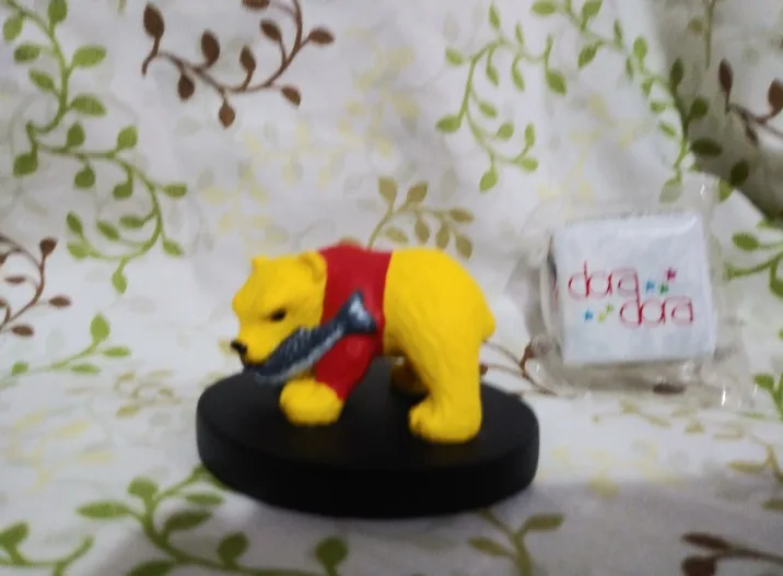 A blurry photo shows a yellow Winnie the Pooh figurine wearing a red shirt and holding a fish, standing on a black base. To…