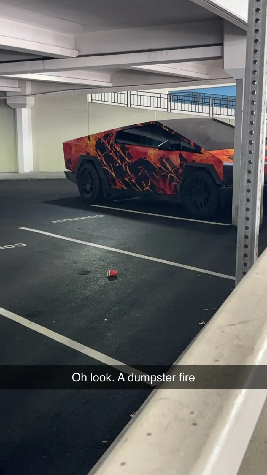 A Tesla Cybertruck with a fiery lava-like wrap is parked in an underground garage, partially obscured by a metal pillar. On…