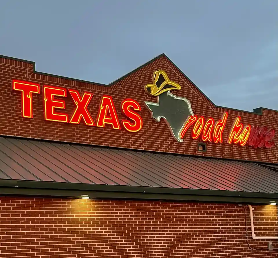 A photo of a Texas Road House restaurant's exterior sign at dusk, featuring red neon letters spelling 'TEXAS' and 'road…