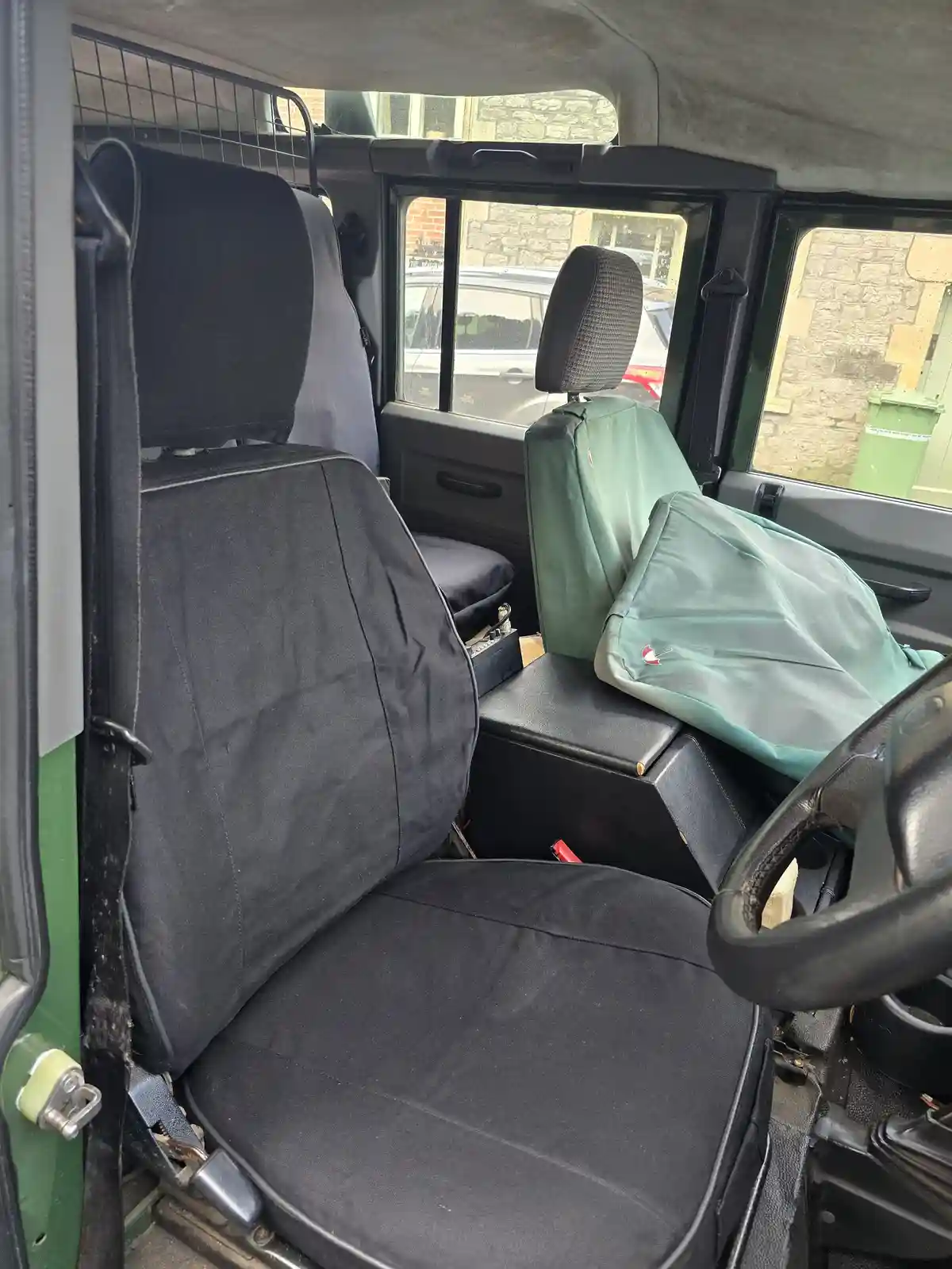 The interior of a green Land Rover Defender shows two seats covered in protective fabric, one black and one green, with the…