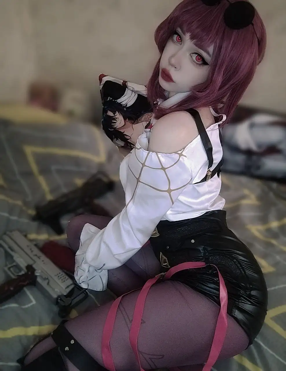 A person in cosplay with purple hair and red eyes sits on a bed holding a pillow with an anime character, surrounded by…