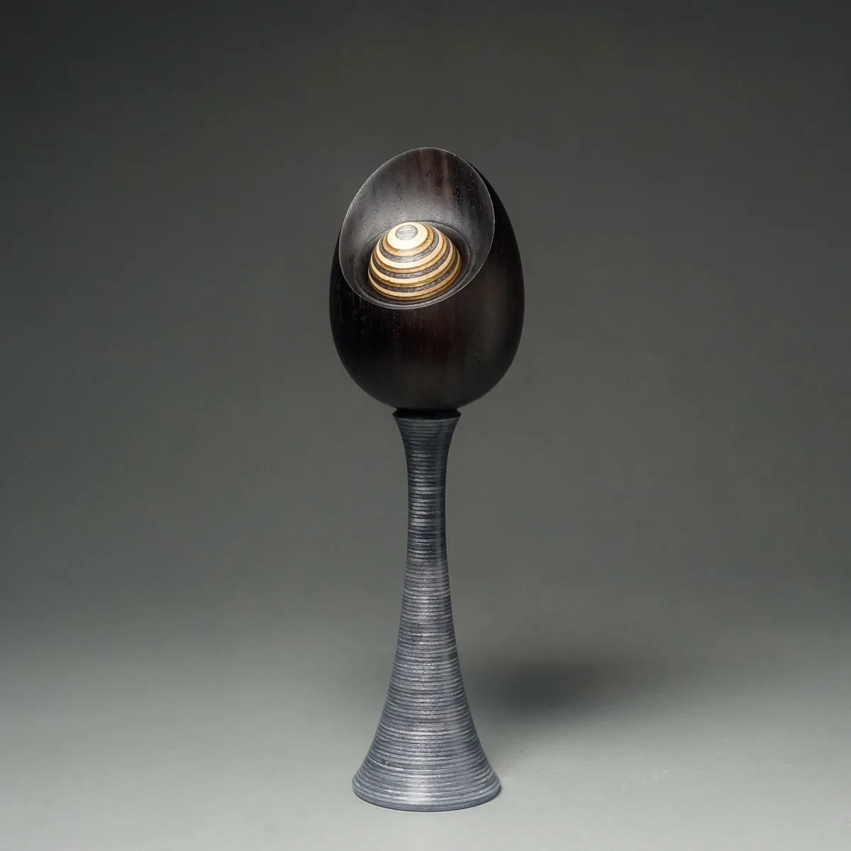 A dark wooden sculpture with a spiraling, striped wooden top sits on a tall, ribbed metallic pedestal against a plain gray…