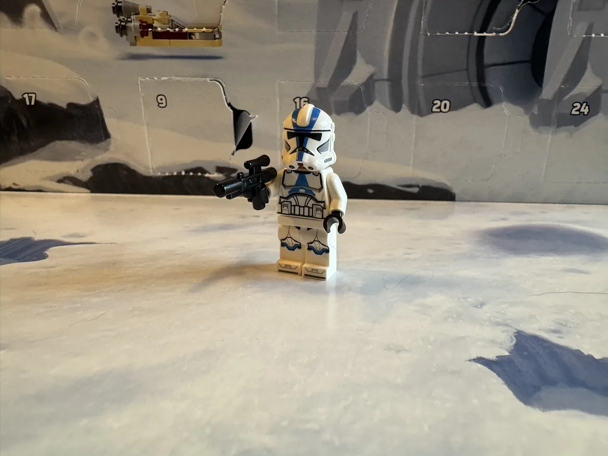 A Lego Star Wars clone trooper minifigure stands on a snowy surface in front of a partially assembled Lego box backdrop with…