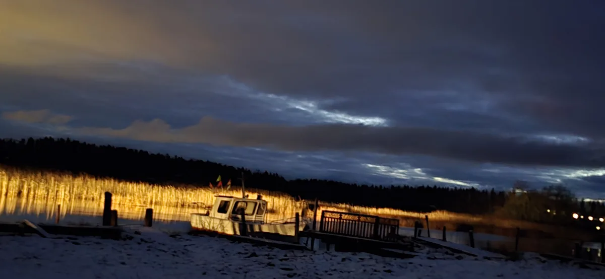 A blurry photo of a boat docked at a snowy waterfront during twilight, with golden light reflecting off the water and dark…