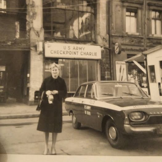 A black-and-white photo shows a woman in a dark coat standing on a street next to a vintage car with 'U.S. ARMY' written on…
