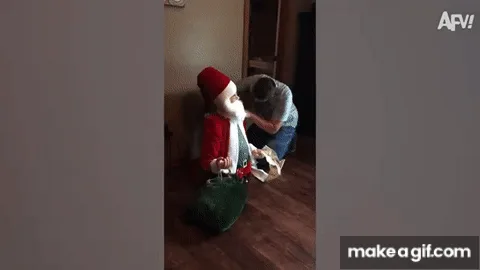 A man in a grey t-shirt kneels beside a Santa Claus costume laid out on the floor, appearing to adjust or fix it. In the…