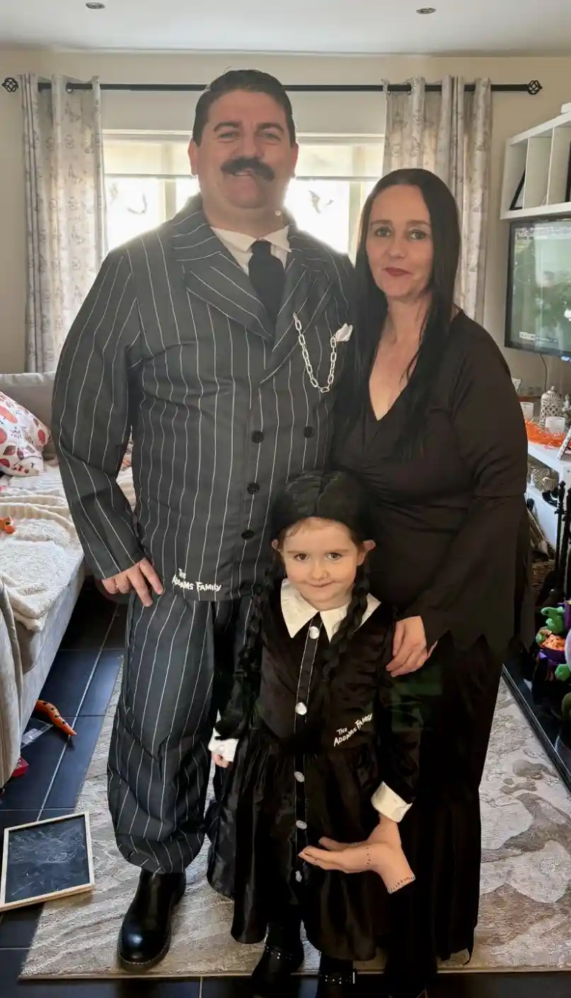 A family of three poses indoors for a photo, dressed in costumes from 'The Addams Family'. The adult male wears a pinstripe…