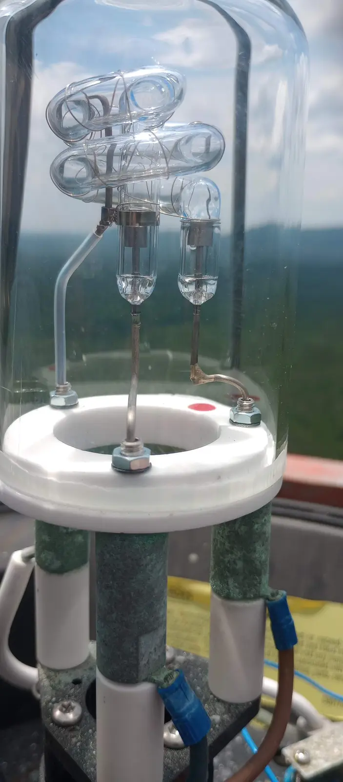 A close-up photo shows a complex, glass-encased scientific apparatus with coiled tubes and metal fittings, mounted on a…