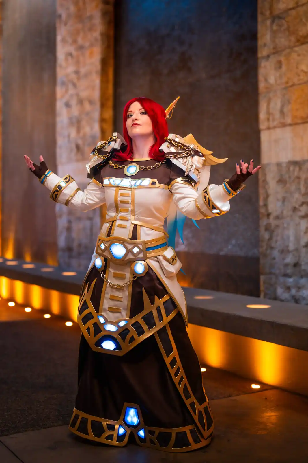 A person in a detailed cosplay of a fantasy character with red hair, white and gold armor, and glowing blue crystals stands…