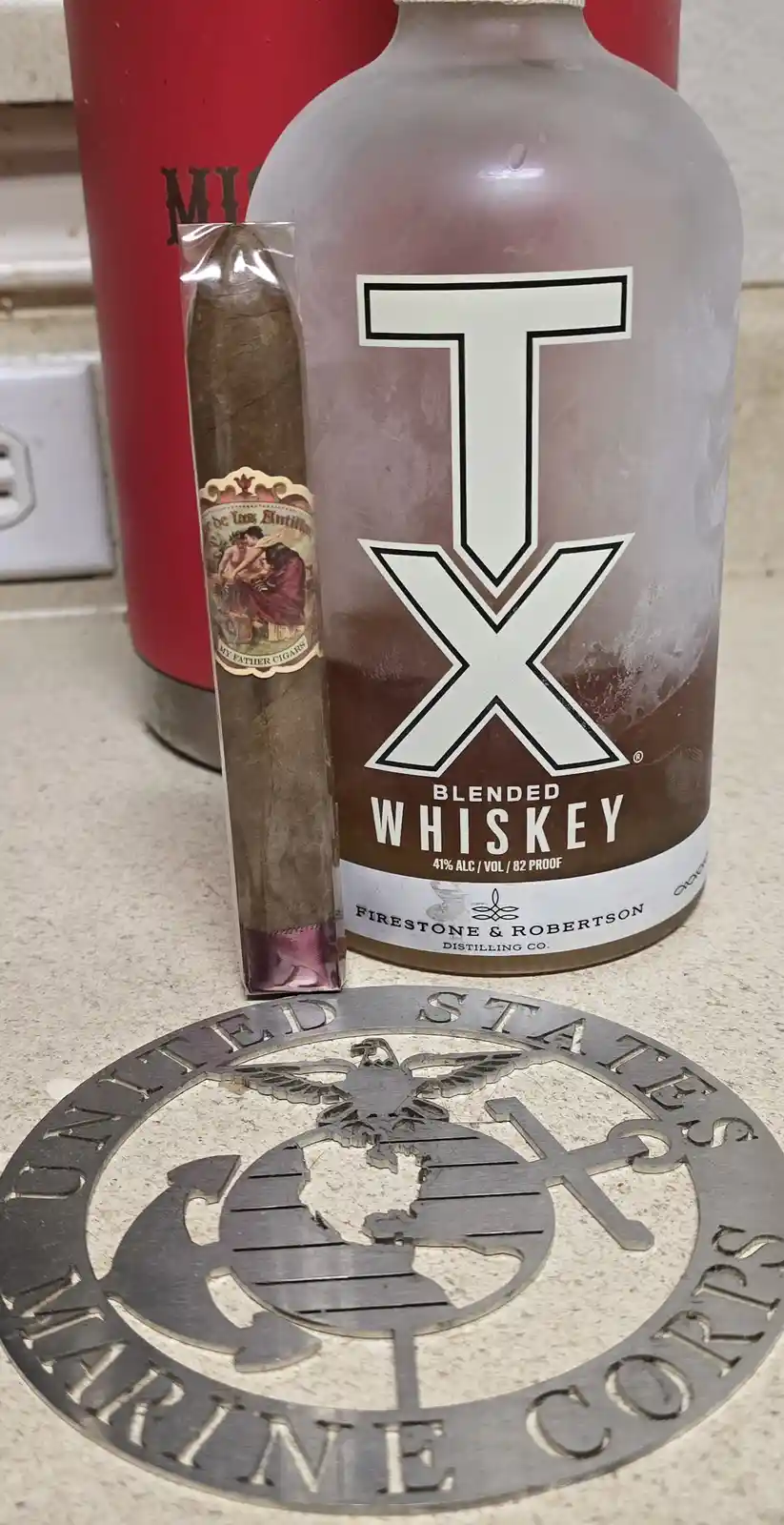 A bottle of TX Blended Whiskey sits next to a My Father Cigars cigar on a countertop, with a United States Marine Corps…