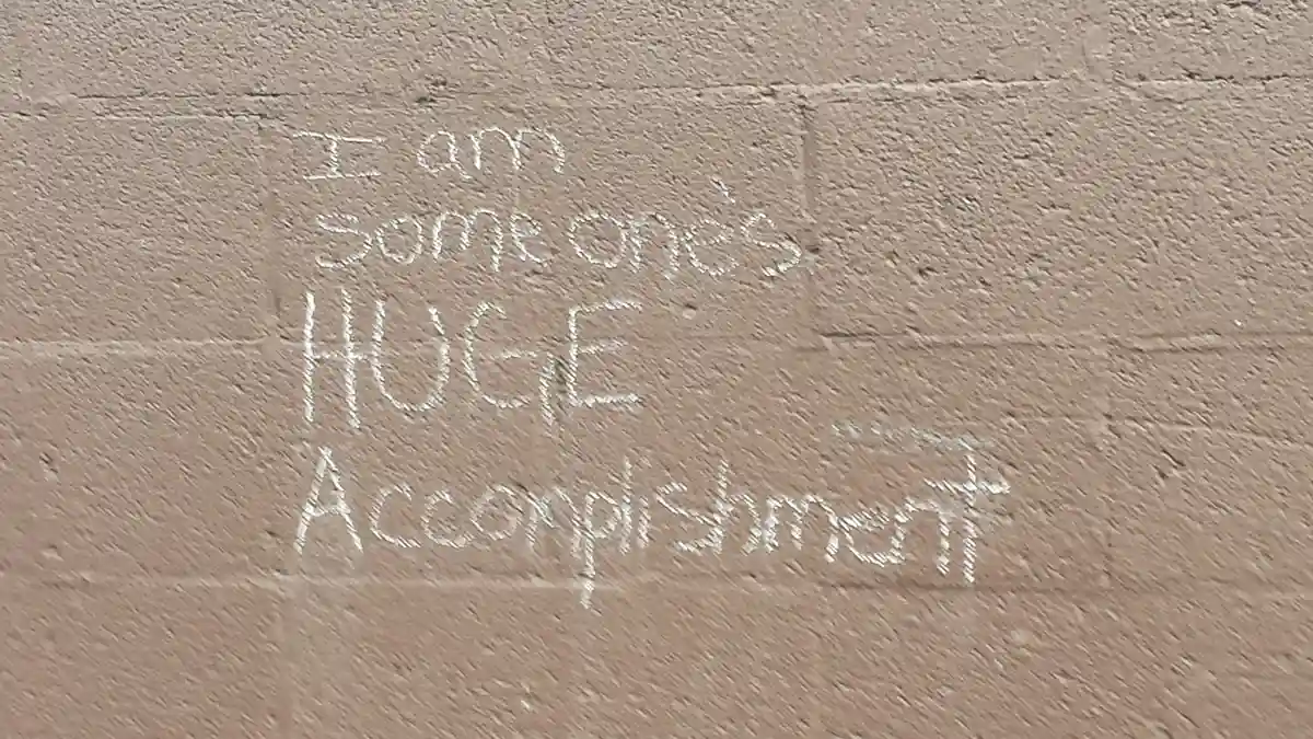 White chalk writing on a beige brick wall reads 'I am someone's HUGE Accomplishment', with 'HUGE' in all caps. The text is…