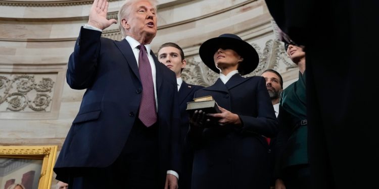 A man in a dark suit and tie raises his right hand while speaking, standing beside a woman in a dark coat and wide-brimmed…