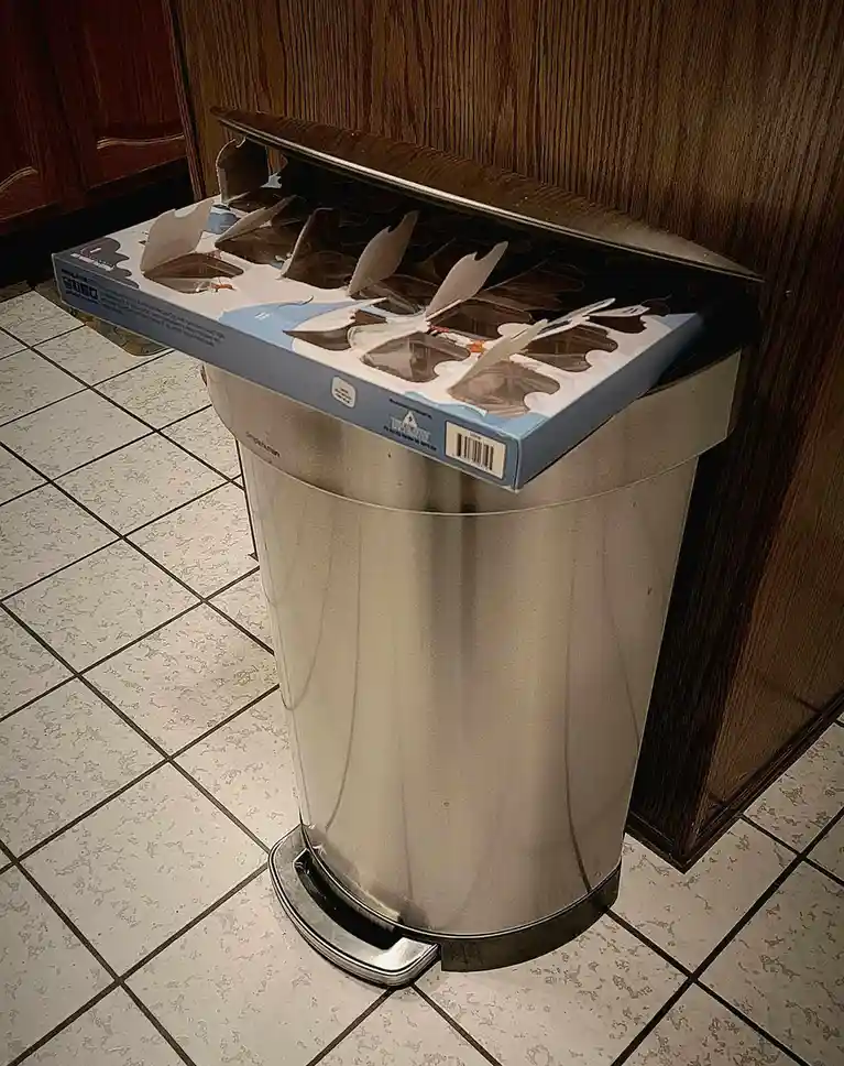 A stainless steel trash can sits on a tiled floor next to wooden cabinets, with an open box of Batman-themed chocolate bars…