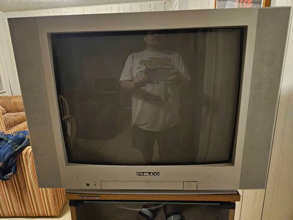 A person wearing a graphic t-shirt and shorts is reflected in the screen of an old, bulky Philco CRT television, holding a…